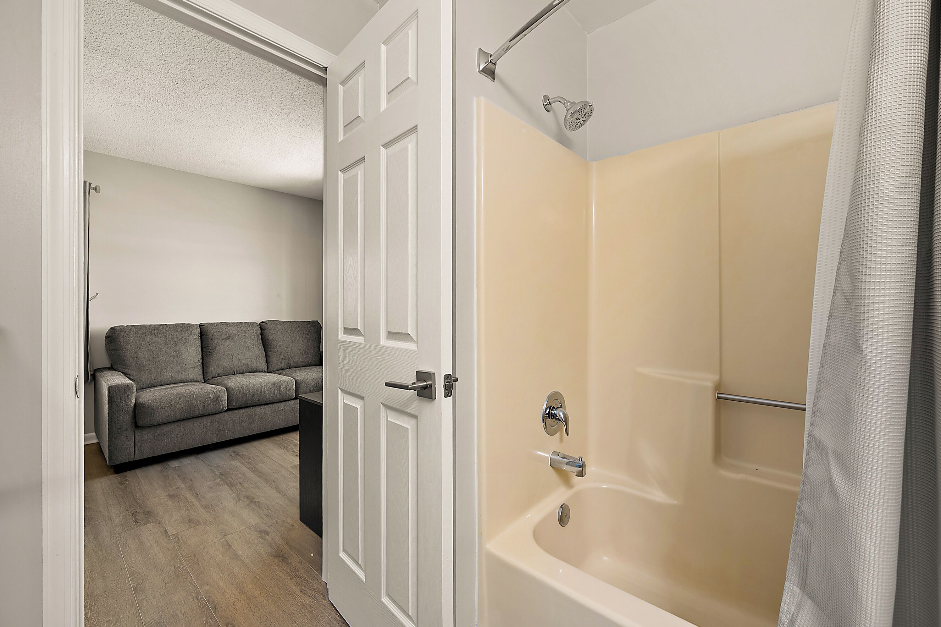 A bathroom with a bathtub, a shower, and a door leading to a living area with a sofa.