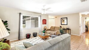 A living room with a sofa, coffee table, and a ceiling fan. There is a window with blinds and a plant on the left. - Refreshing Oasis with Massage Chair, Game Garage, Large Fenced Yard +Retro games (Longwood)