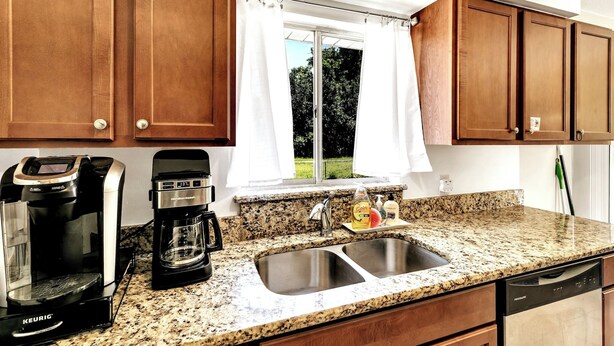 A kitchen with granite countertops, two coffee makers, a sink, wooden cabinets, and a window with white curtains. - Refreshing Oasis with Massage Chair, Game Garage, Large Fenced Yard +Retro games (Longwood)