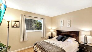 A bedroom with a bed, bedside tables, a window with curtains, a wall-mounted TV, and framed pictures. - Refreshing Oasis with Massage Chair, Game Garage, Large Fenced Yard +Retro games (Longwood)