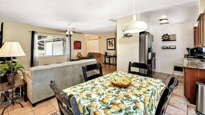 A dining area with a table covered in a fruit pattern tablecloth, a ceiling fan, and a kitchen area with a refrigerator and cabinets. - Refreshing Oasis with Massage Chair, Game Garage, Large Fenced Yard +Retro games (Longwood)