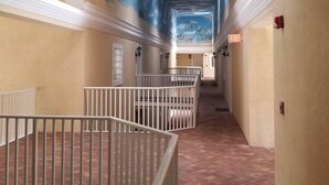 Ein Hotelkorridor mit Fliesenboden, weißen Geländern und einem Wandgemälde an der Decke, das eine tropische Szene zeigt.