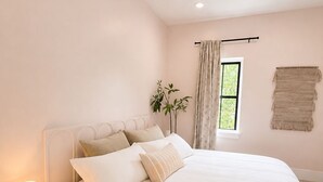 Un dormitorio con cama, lámpara de noche, ventana con cortinas, una planta y un macramé colgado en la pared.