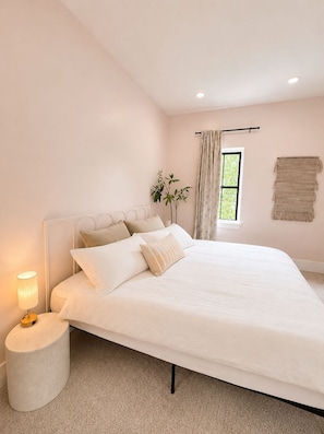 A bedroom with a bed, a bedside lamp, a window with curtains, a plant, and a wall-mounted macrame.