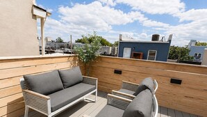 Un patio en la azotea con sofá y sillones, una terraza de madera y un cielo azul despejado.