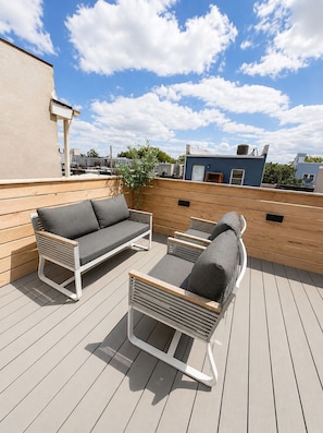 A rooftop patio with a sofa and chairs, a wooden deck, and a clear blue sky.