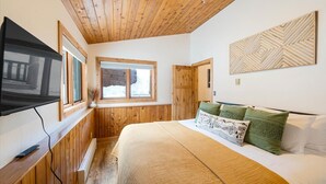 A bedroom with wooden paneling, a bed with green and white pillows, a mounted TV, and a window with a view of a snowy landscape. - New Listing! Three Bedroom Apartment, Sleeps 6, Steps to Ski Access (Whitefish)