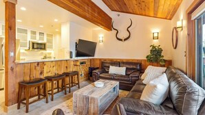 A cozy living room with a wooden ceiling, a leather sofa, a coffee table, and a kitchen area with a flat-screen TV. - New Listing! Three Bedroom Apartment, Sleeps 6, Steps to Ski Access (Whitefish)