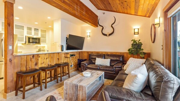 Un salon chaleureux avec un plafond en bois, un canapé en cuir, une table basse et un coin cuisine équipé d’un téléviseur à écran plat.