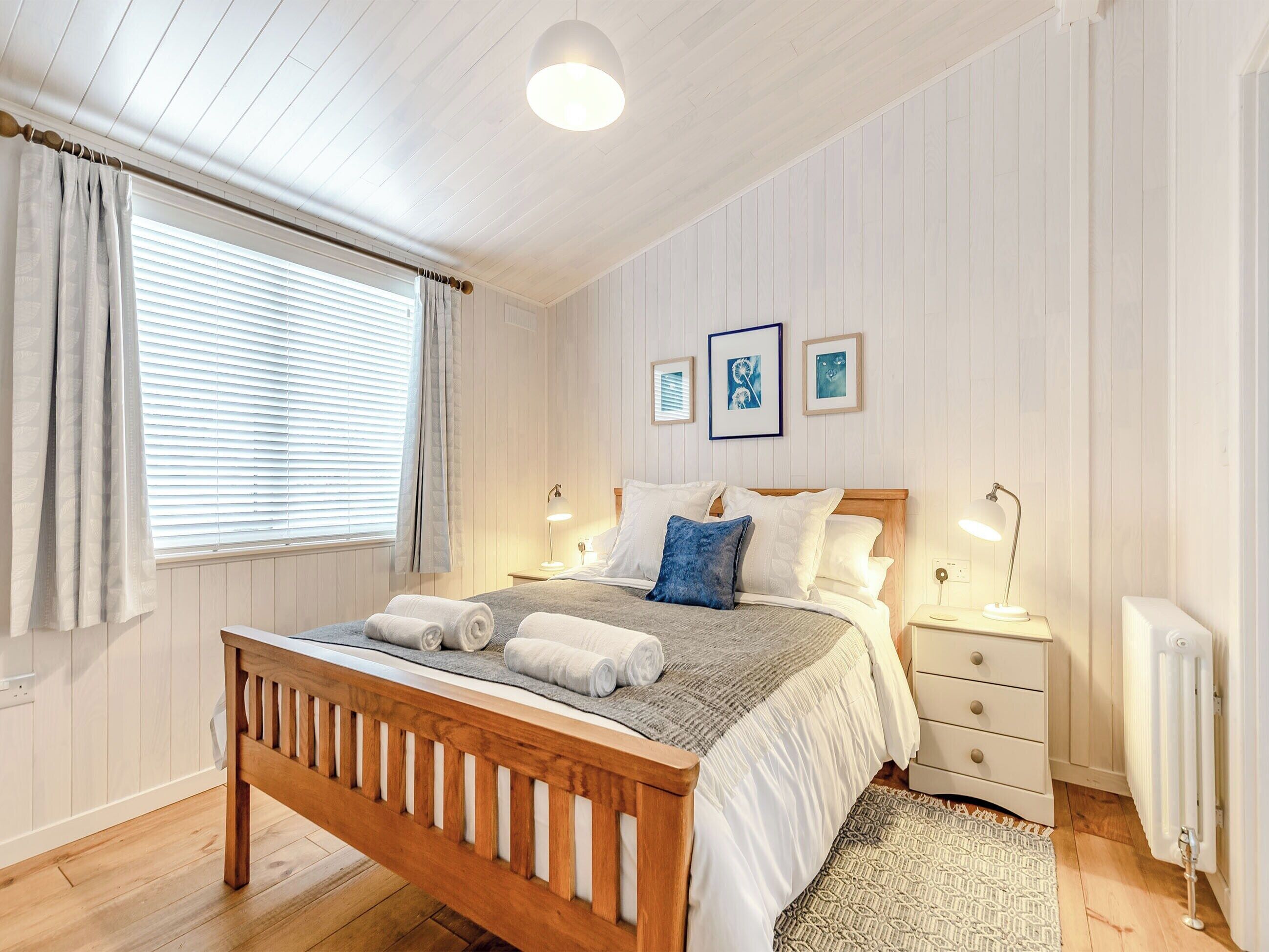 A bedroom with a wooden bed, white bedding, two bedside lamps, a radiator, and a window with curtains.