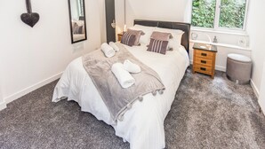 Ein ordentlich bezogenes Bett mit grauer Decke und Kissen, ein Nachttisch mit Lampe, einem Spiegel und ein Fenster mit Blick auf Grün.