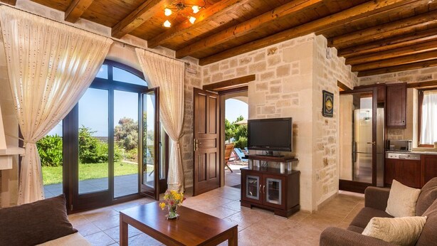 Un salón con techo de madera, paredes de piedra, un televisor sobre un mueble y vistas al exterior.