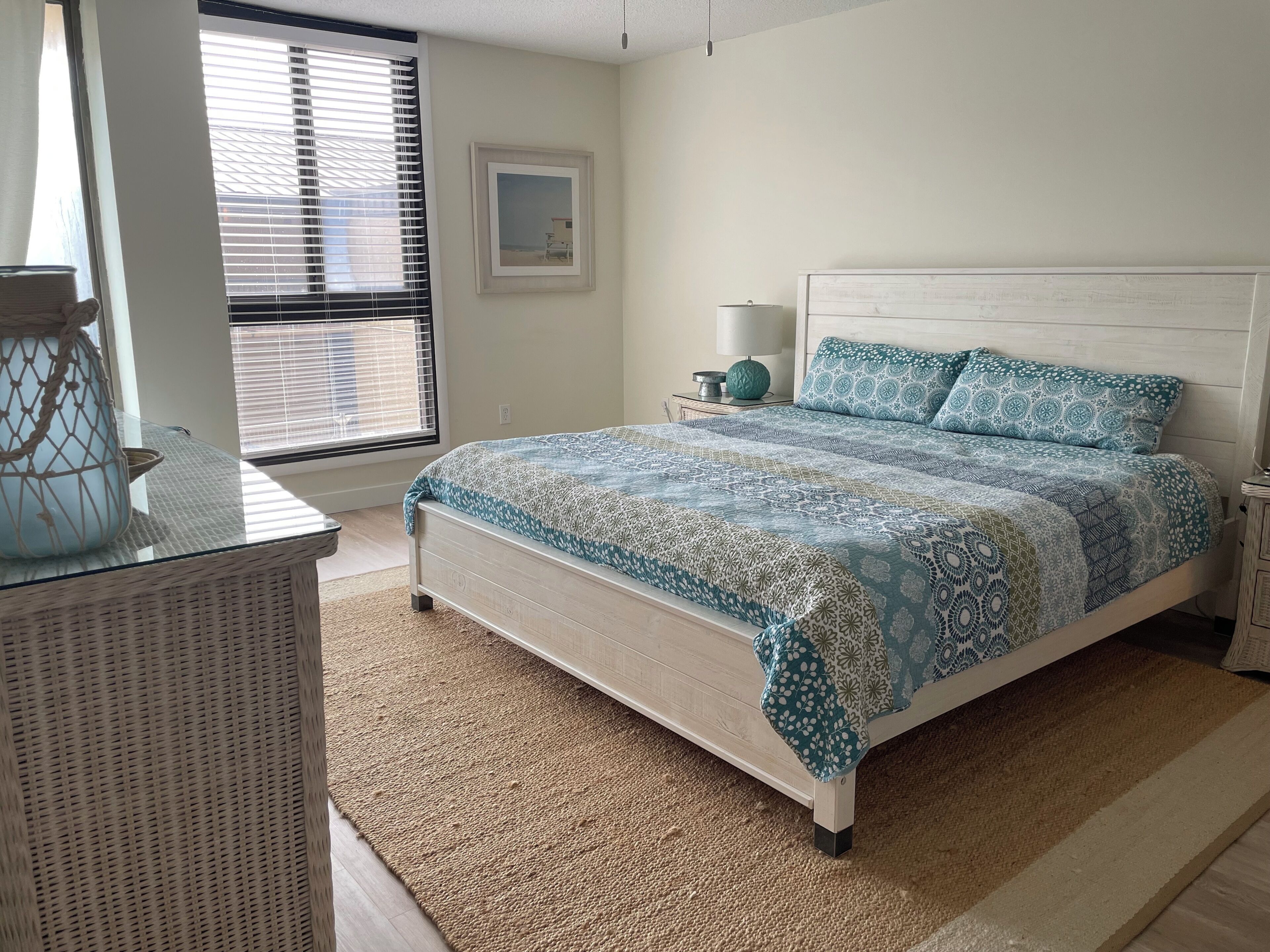 A bedroom with a bed, a nightstand, a lamp, a picture on the wall, and a window with blinds.