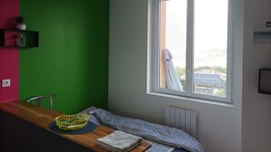 A room with a green wall, a wooden table, a window with a view of solar panels, and a radiator. - Studio Welcome to Keylia (Saint-Étienne)