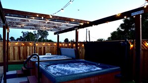 A hot tub under a wooden pergola with string lights, surrounded by a wooden fence and steps leading up to it. - Welcome to Oasis On The Links! (Paso Robles)