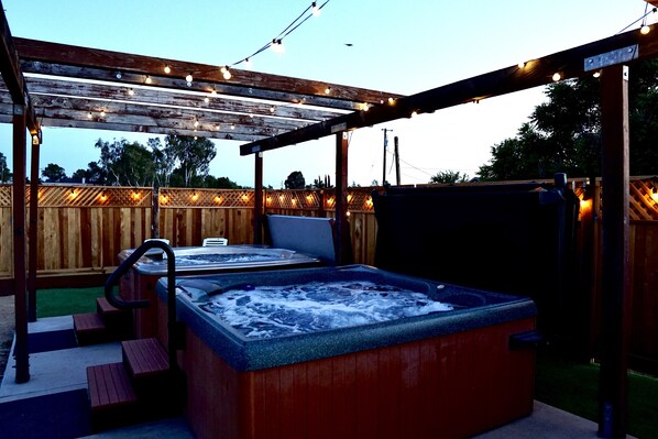 A hot tub under a wooden pergola with string lights, surrounded by a wooden fence and steps leading up to it.