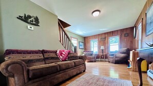 A spacious living room with a brown leather couch, a patterned rug, wooden floors, a staircase, and a piano. - The Guides House • Cozy Main St Stay with Porch Views (Old Forge)