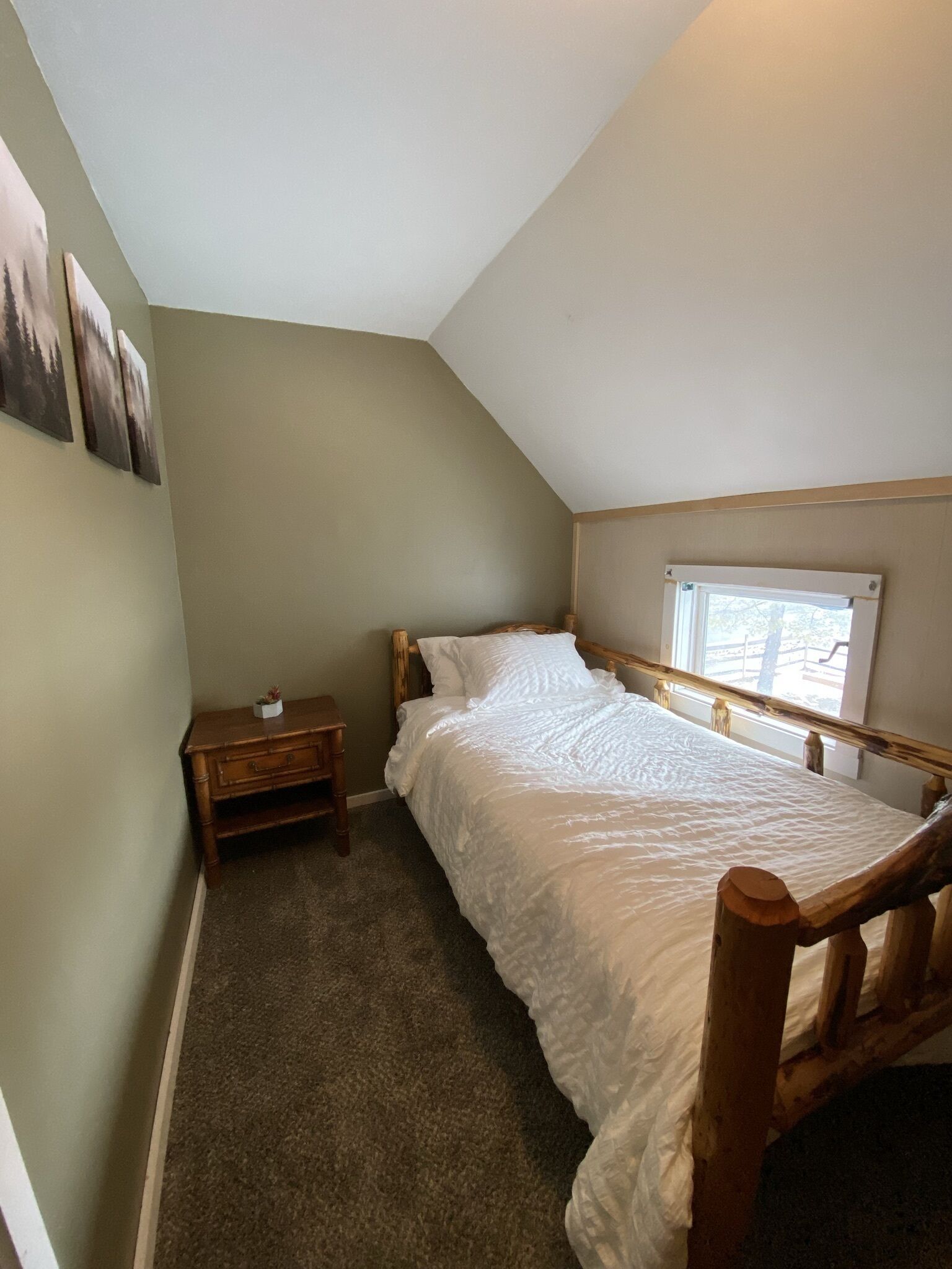 A small, single-bed room with a sloped ceiling, a wooden nightstand, a window with a view, and a wall-mounted shelf.