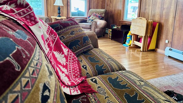 A cozy living room with wooden walls, a sofa with patterned blankets, a side table with a lamp, and a window with a view of trees. - The Guides House • Cozy Main St Stay with Porch Views (Old Forge)