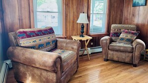 A cozy room with wooden walls and floors, featuring two leather armchairs with patterned cushions, a small wooden table with a lamp, and large windows with blinds. - The Guides House • Cozy Main St Stay with Porch Views (Old Forge)