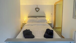 Un lit avec du linge de lit blanc, deux lampes de chevet, une décoration en forme de cœur accrochée au mur et un miroir sur le mur.