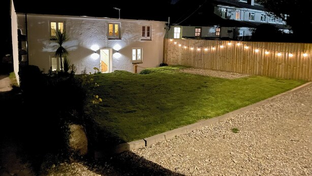 Une maison dotée d’un éclairage extérieur soigné, d’une cour verdoyante et d’une clôture en bois.