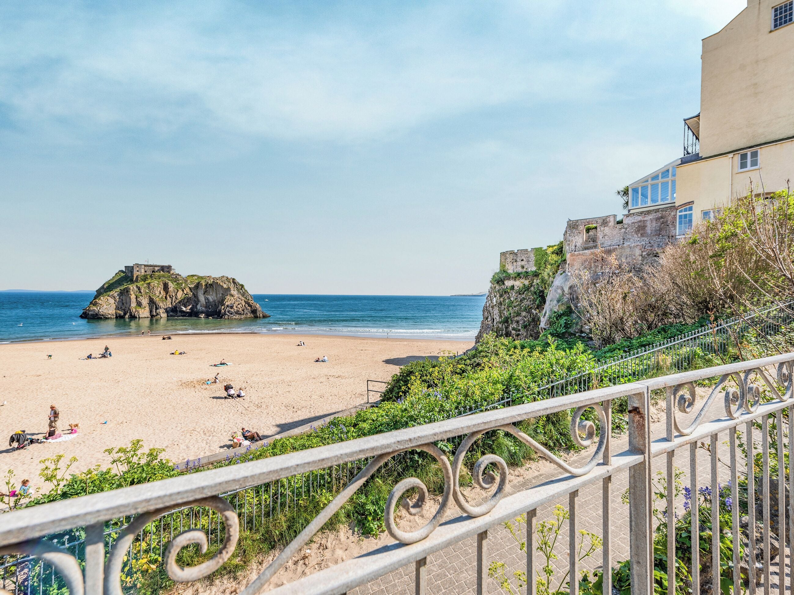 Una playa con una estructura de piedra, un edificio con balcón y una barandilla de metal.