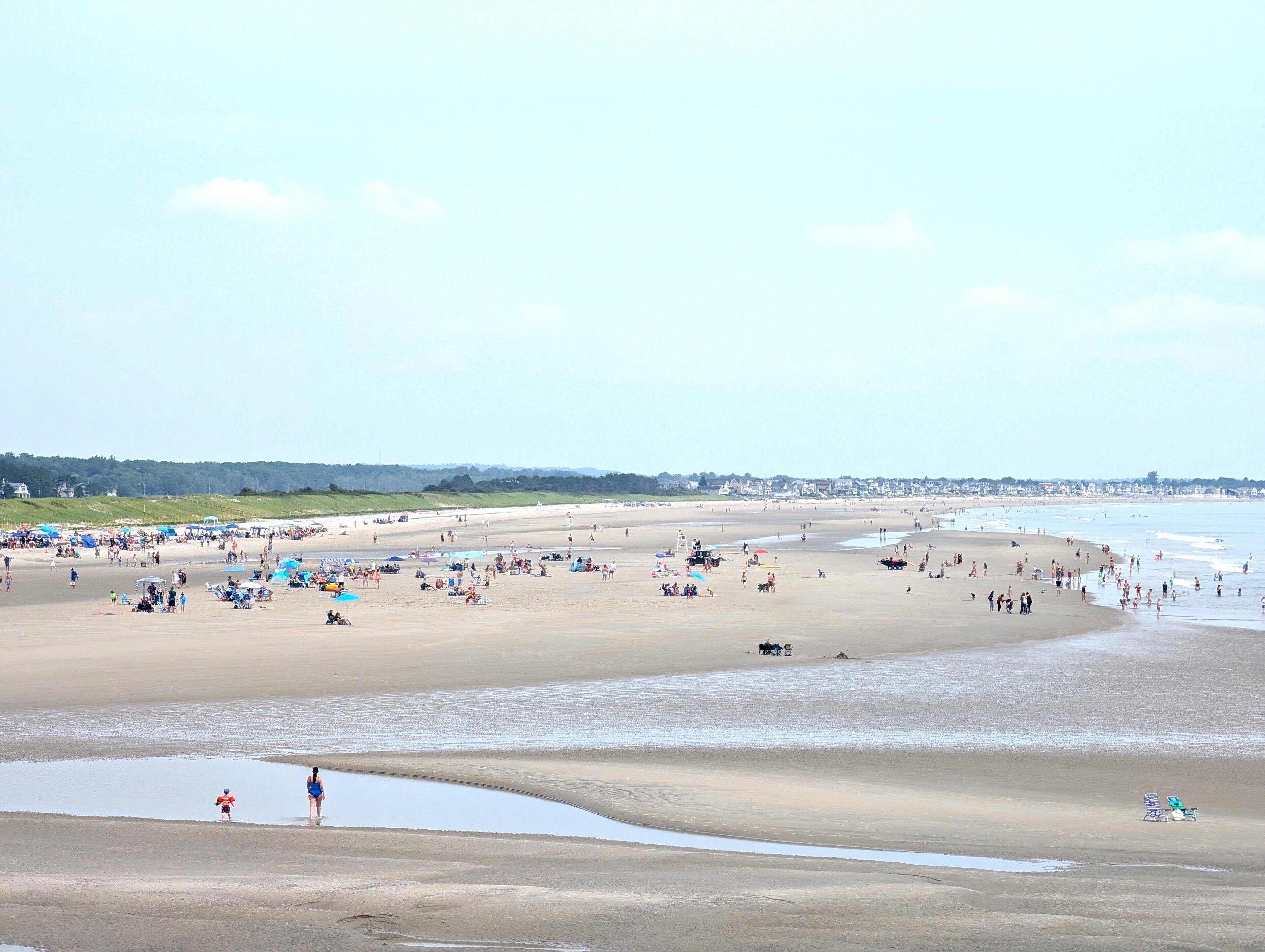 Une plage avec des gens et des parasols, un ciel dégagé et une côte lointaine.