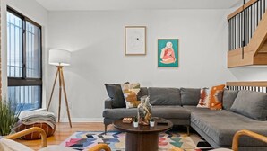 A modern living room with a grey sectional sofa, a round coffee table, and a floor lamp. There are two framed artworks on the wall and a staircase to the right.