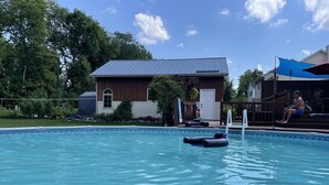 Una piscina con una casa de madera al fondo, un espacio cubierto donde se sienta una persona, y un cielo azul despejado.