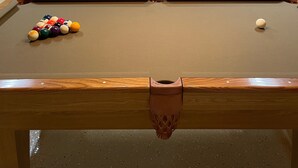 Una mesa de billar con bolas, una mesa de comedor con sillas, una estufa y un microondas en una habitación con paredes de madera y una lámpara colgante.