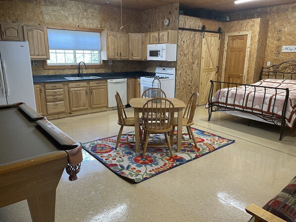 A compact kitchen with wooden cabinets, a dining table with chairs, a microwave, and a bed with a metal frame.