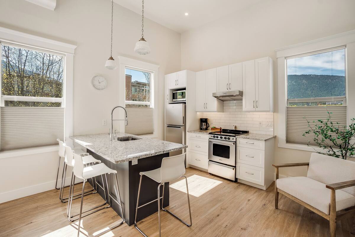 Una cocina moderna con una isla central, muebles blancos, electrodomésticos de acero inoxidable y ventanas amplias que ofrecen vistas al exterior.