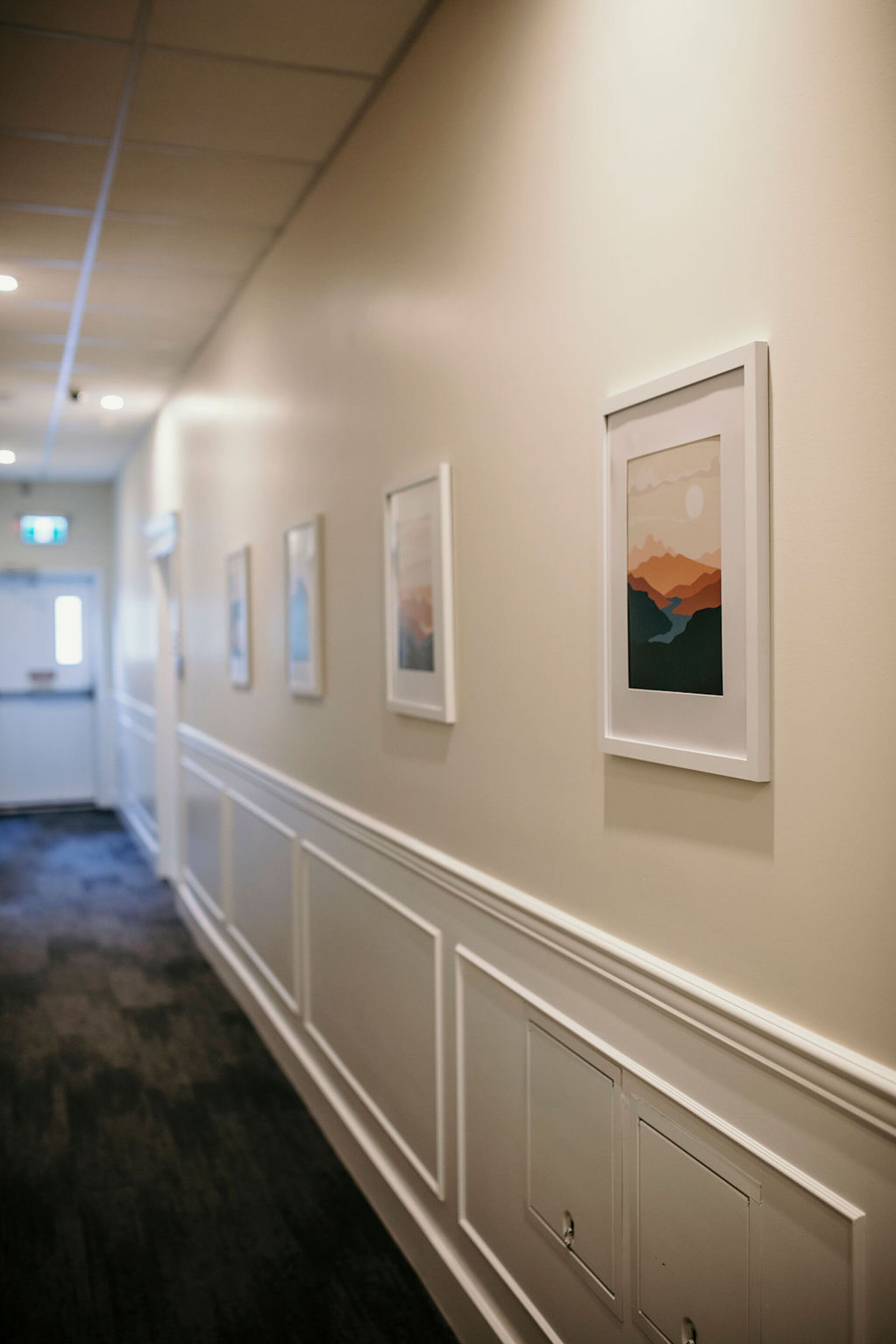 A hallway with framed artwork on the walls, a carpeted floor, and a door at the end.