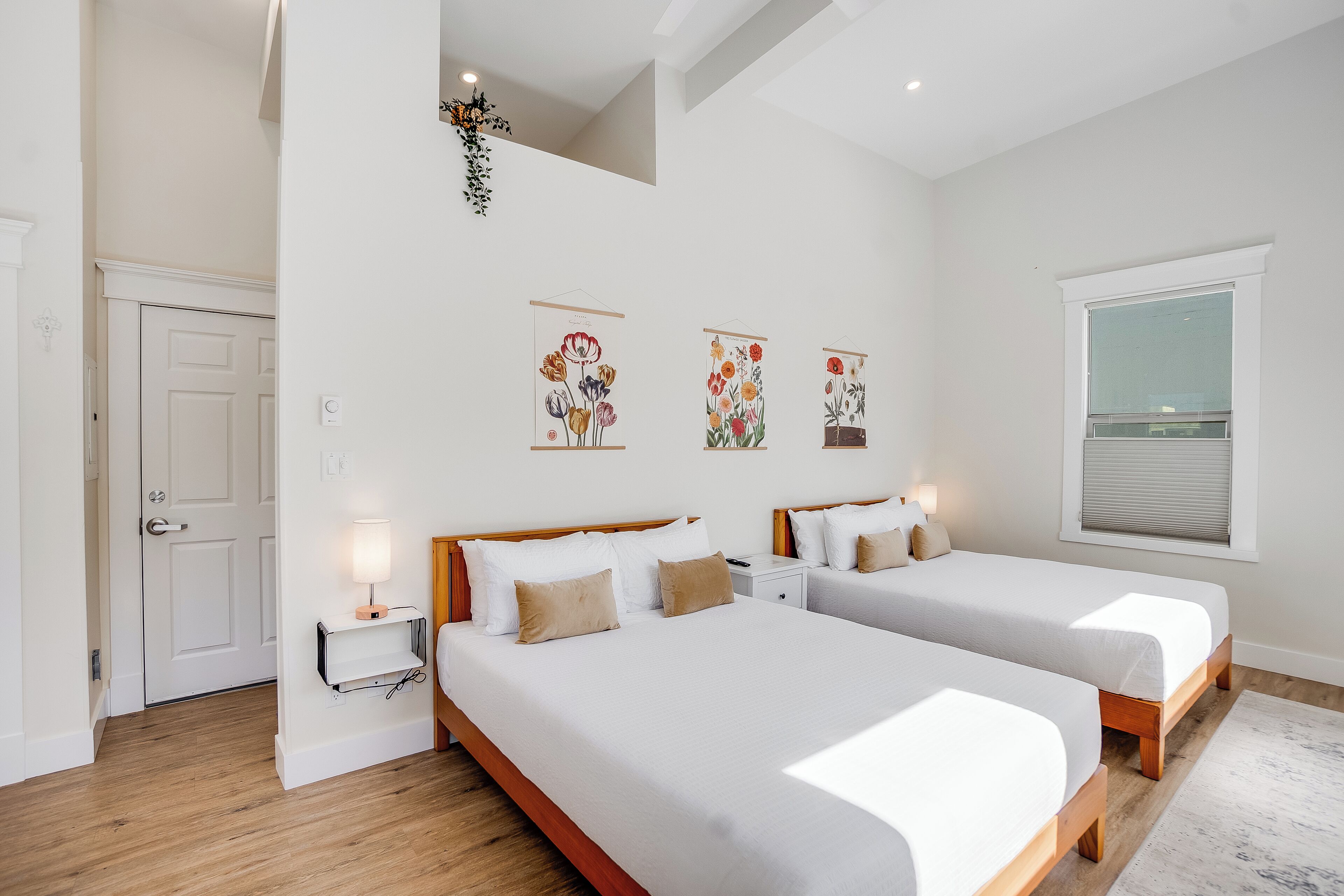 A bedroom with two beds, a white door, a small bedside table with a lamp, and wall art.