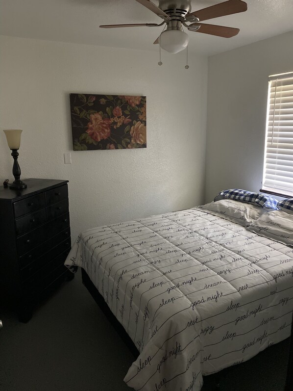 A bedroom with a bed, a dresser, a ceiling fan, a window with blinds, and a floral painting on the wall.