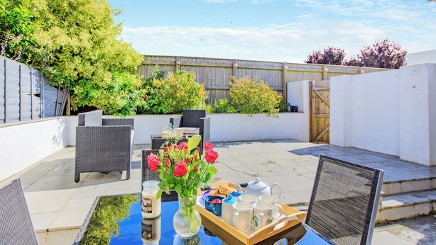 Une terrasse extérieure avec une table en verre, des chaises en osier, un vase de roses rouges et un plateau avec des tasses et une théière.