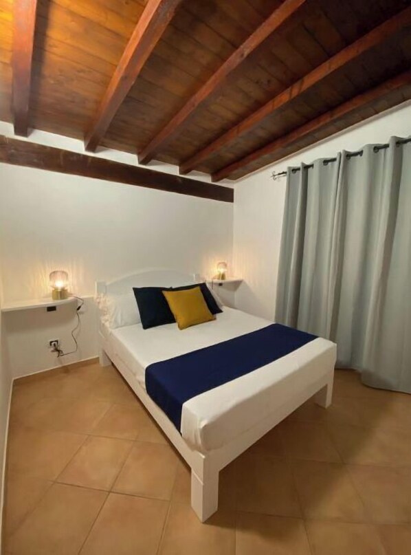 Un dormitorio con techo de madera, una cama con colcha azul, dos lámparas de noche y un ventanal con cortinas.