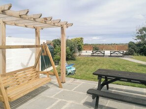 Una pérgola de madera, un columpio, una mesa de picnic y un patio cubierto de césped con una cerca blanca y un muro de ladrillo al fondo.