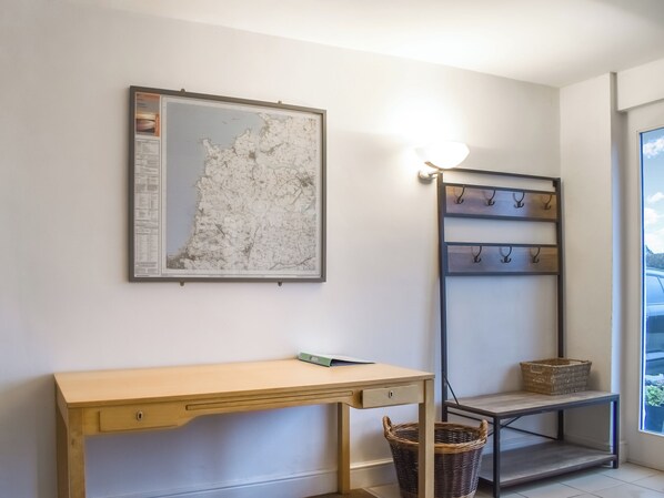 A room with a wooden desk, a framed map on the wall, a wall-mounted light, a shelf with hooks, and a basket.