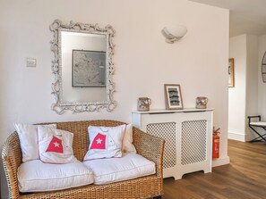 Ein Korbusofa mit weißen und roten Kissen, ein weißes Schränkchen mit verzierten Türen, ein großer, aufwendiger Spiegel und ein gerahmtes Bild an der Wand.
