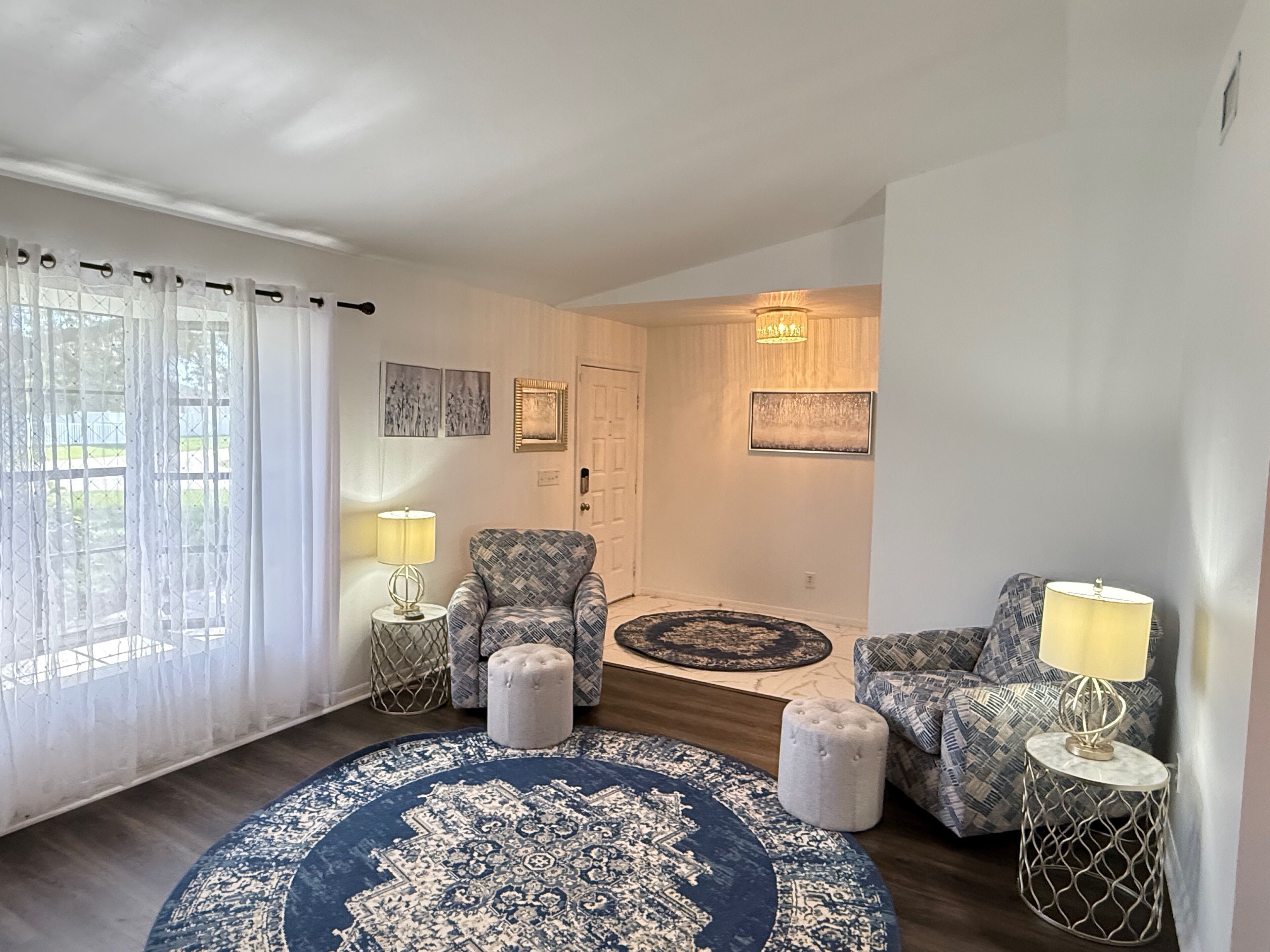 Ein Wohnzimmer mit einem großen, blau gemusterten Teppich, zwei Sesseln, einem kleinen Hocker und einem runden Couchtisch. Zwei Lampen stehen auf Beistelltischen, eine Tür mit einer Leuchte darüber und Kunstwerken an den Wänden.