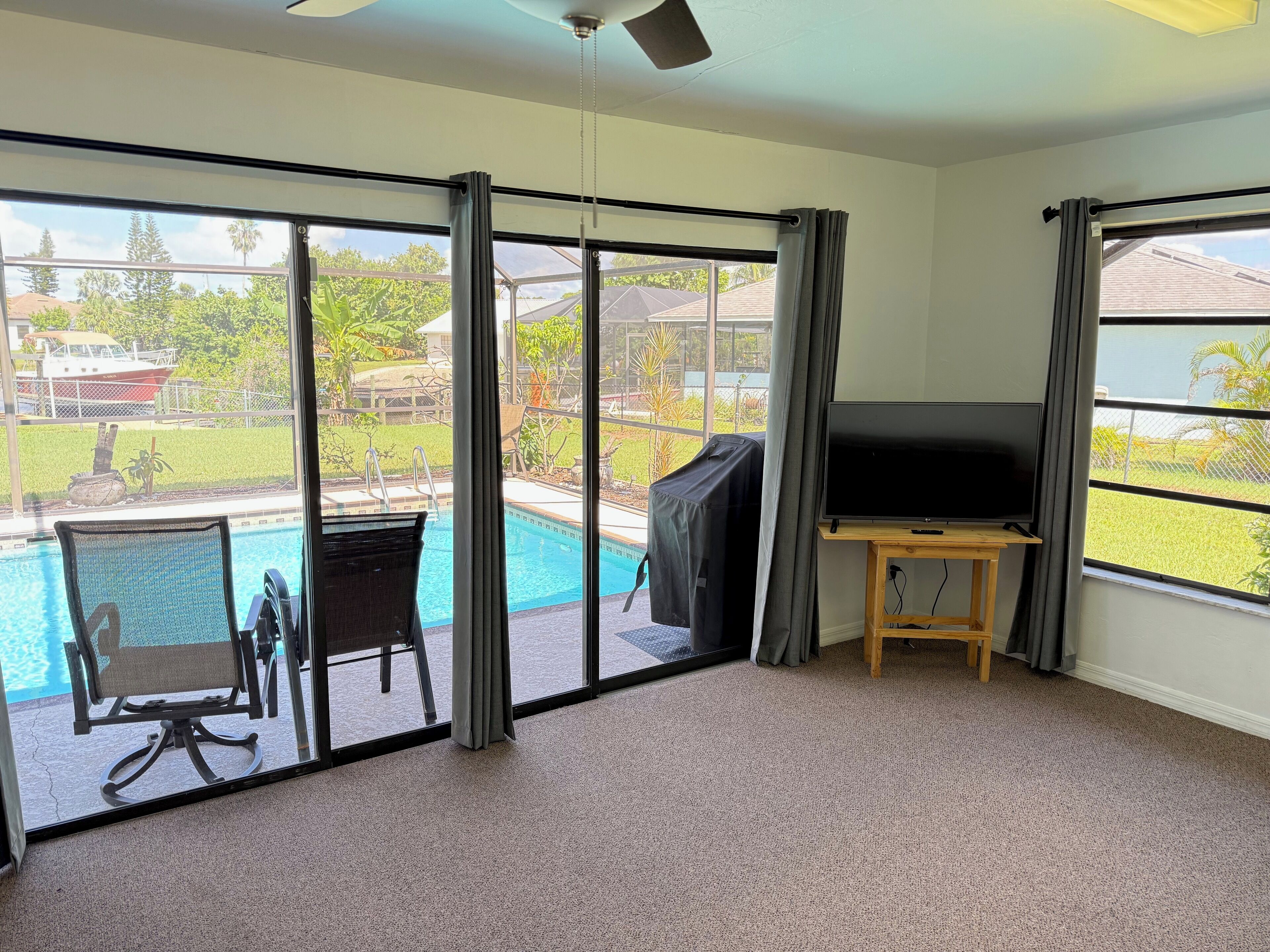 A room with a large sliding glass door leading to a pool area, a ceiling fan, and a flat-screen TV on a wooden stand.