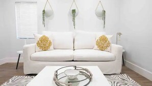 Een witte bank met kussens met goudkleurige patronen, een glazen salontafel met een decoratieve metalen dienblad, en hangende groene planten.