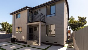 A modern two-story house with a balcony, a staircase leading to the entrance, and a small outdoor area with artificial grass.