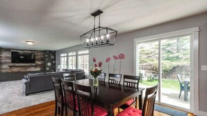 A dining area with a wooden table and chairs, a chandelier, and a view of the backyard through large windows. - Upscale House for up to 20 with Fire Pit & Pool (Meridian)