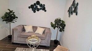 Un salon avec un canapé gris, deux plantes en pot, une petite table basse ronde en verre et un petit tabouret.