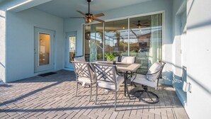 Une terrasse couverte avec une table et des chaises, un ventilateur de plafond et une porte vitrée donnant sur un balcon.