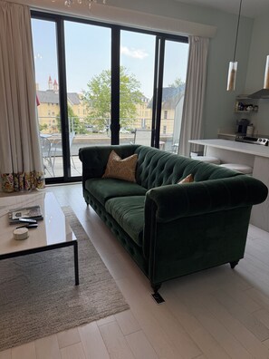A green sofa with a cushion, a coffee table with books and a remote, and a view of buildings through large windows. - Asylum 58 Condo @ Grand Traverse Commons (Traverse City)