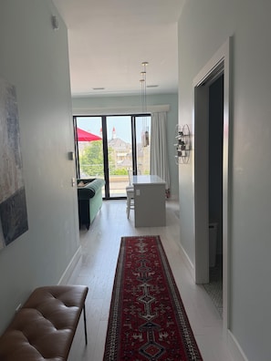 Un intérieur moderne avec un banc, un tapis rouge à motifs et une vue, à travers une porte vitrée, sur un espace extérieur agrémenté d’un parasol rouge.
